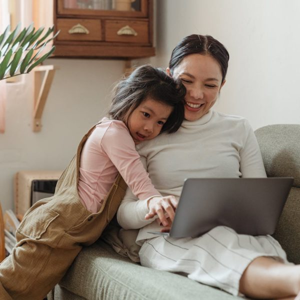 Cheerful smiling Asian woman browsing modern netbook while hugging with cute content daughter on comfy sofa in cozy living room
